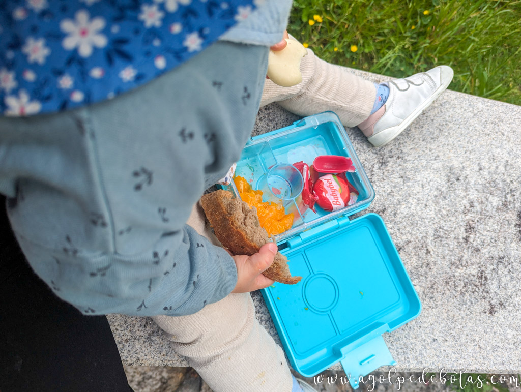 Niña pequeña comiendo pan con queso y mandarina durante un picnic en la montaña, sentada sobre una piedra con una fiambrera azul.